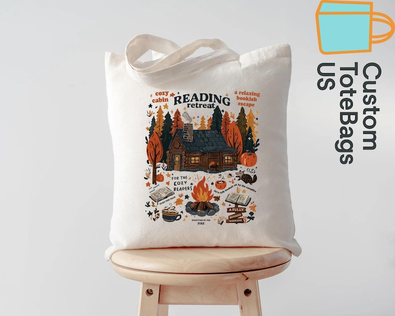 May include: Cream-coloured tote bag with a colourful illustration of a cabin in a forest, a campfire, and books. Text on the bag reads "Reading Retreat" and "For the cozy readers". The bag has a simple design with two handles.