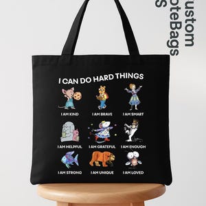May include: Black tote bag with the text "I CAN DO HARD THINGS" and illustrations of various characters with positive affirmations like "I AM KIND" and "I AM BRAVE". The bag has black handles.