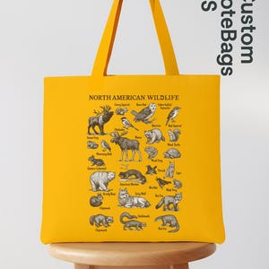 May include: A mustard yellow tote bag displaying a detailed illustration of North American wildlife. The bag features various animals and birds, with the text "NORTH AMERICAN WILDLIFE". The bag also has the text "Custom ToteBags US".