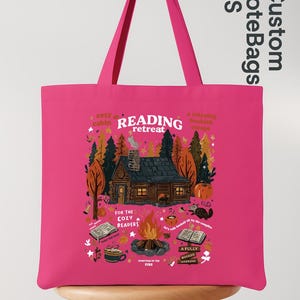 May include: A vibrant pink tote bag with a cabin scene and the words "READING retreat". The design includes a cabin, a campfire, books, and the text "For the Cozy Readers". The bag has long handles.
