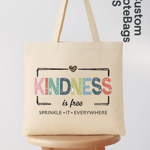 May include: Cream-colored tote bag with the words "KINDNESS is free" in colorful letters. The bag has a black rectangular border and a leopard print heart. The phrase "SPRINKLE &bull; IT &bull; EVERYWHERE" is printed below.