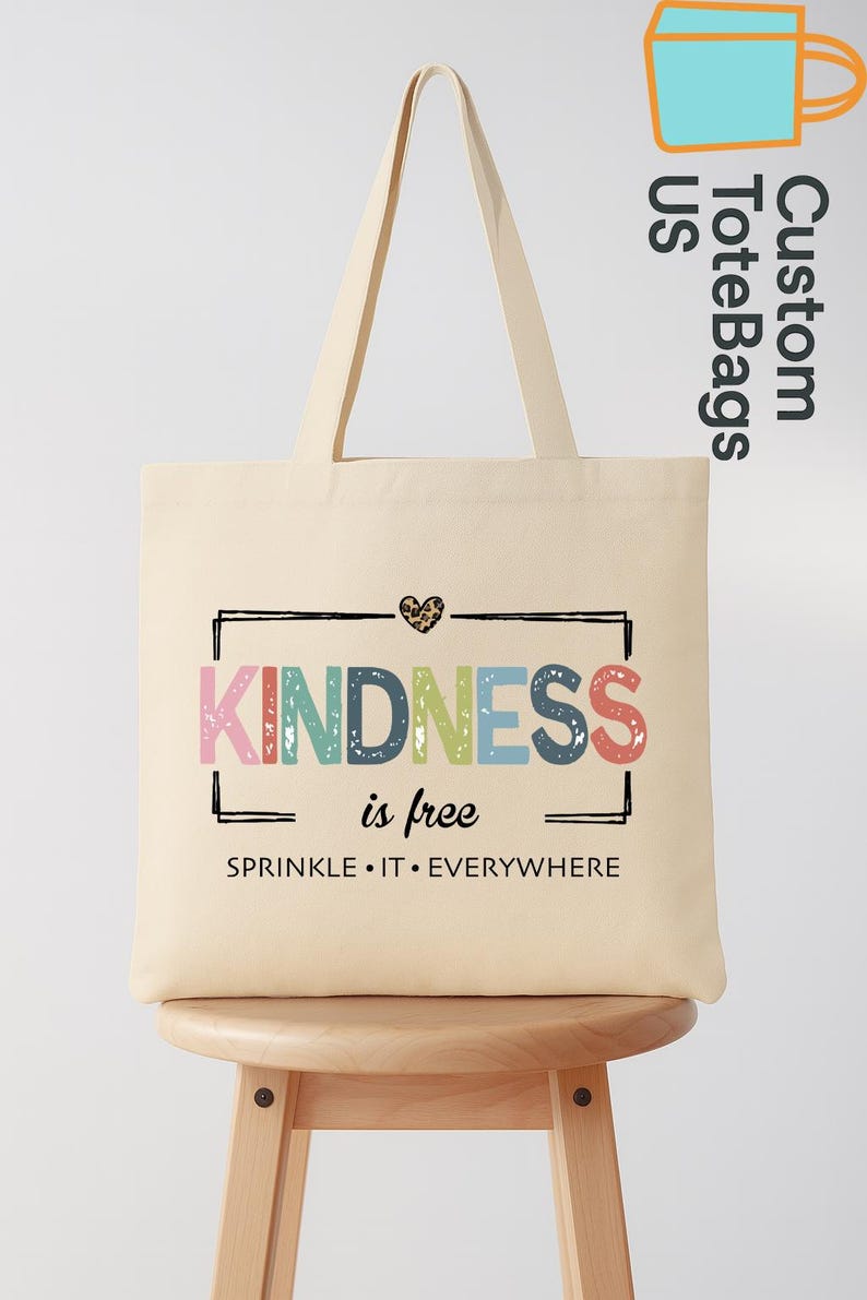 May include: Cream-colored tote bag with the words "KINDNESS is free" in colorful letters. The bag has a black rectangular border and a leopard print heart. The phrase "SPRINKLE &bull; IT &bull; EVERYWHERE" is printed below.