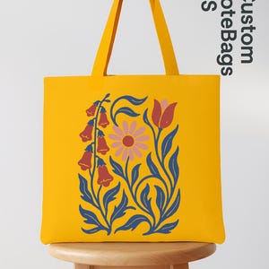 Peut inclure: Un sac fourre-tout jaune avec un motif floral. Le sac pr&eacute;sente un motif stylis&eacute; de fleurs et de feuilles en bleu, rose et rouge. Le sac est pos&eacute; sur un tabouret en bois. Le texte "Custom Tote Bags US" est visible.