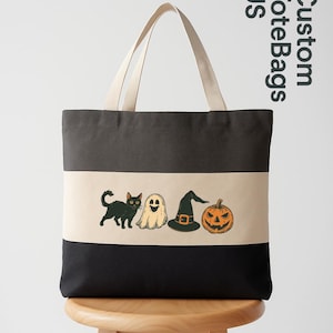 May include: A black, gray, and beige tote bag with a Halloween-themed design. The design includes a black cat, a ghost, a witch's hat, and a jack-o'-lantern. The bag has beige handles and the text "Custom Tote Bags US".