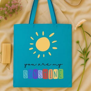 Peut inclure: Un sac fourre-tout turquoise avec des poign&eacute;es bleues, orn&eacute; d'une illustration de soleil jaune et du texte "you are my sunshine" dans une police color&eacute;e et ludique. Un appareil photo et une fleur sont en arri&egrave;re-plan.