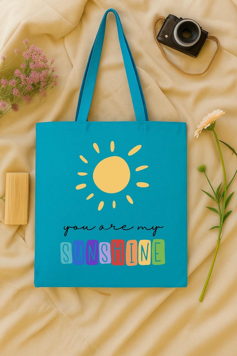 Peut inclure: Un sac fourre-tout turquoise avec des poign&eacute;es bleues, orn&eacute; d'une illustration de soleil jaune et du texte "you are my sunshine" dans une police color&eacute;e et ludique. Un appareil photo et une fleur sont en arri&egrave;re-plan.