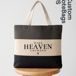 May include: A canvas tote bag with a black, tan, and dark gray striped design. The bag features the text "MAKE HEAVEN CROWDED" with a cross symbol. The tote bag has tan handles and is displayed on a wooden stool.