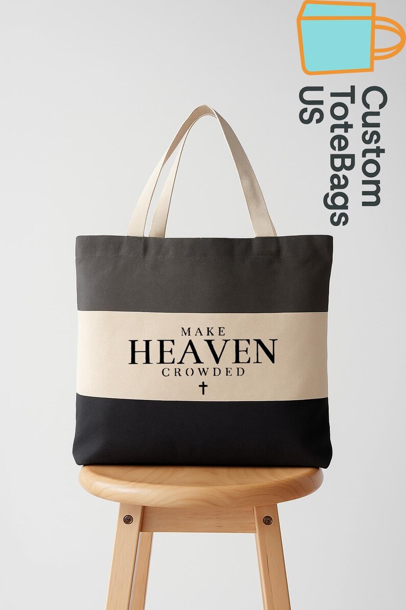 May include: A canvas tote bag with a black, tan, and dark gray striped design. The bag features the text "MAKE HEAVEN CROWDED" with a cross symbol. The tote bag has tan handles and is displayed on a wooden stool.