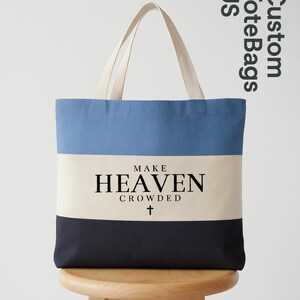 May include: A canvas tote bag with a blue, cream, and navy color block design. The bag features the text "MAKE HEAVEN CROWDED" in black, with a small cross below. The handles are a natural cream color.