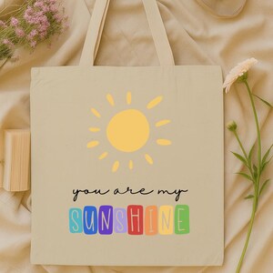 Peut inclure: Sac fourre-tout beige avec un soleil jaune et le texte "you are my sunshine" en lettres color&eacute;es. Le sac a de longues anses et est fait d'un mat&eacute;riau semblable &agrave; de la toile. Un appareil photo noir et des fleurs sont en arri&egrave;re-plan.
