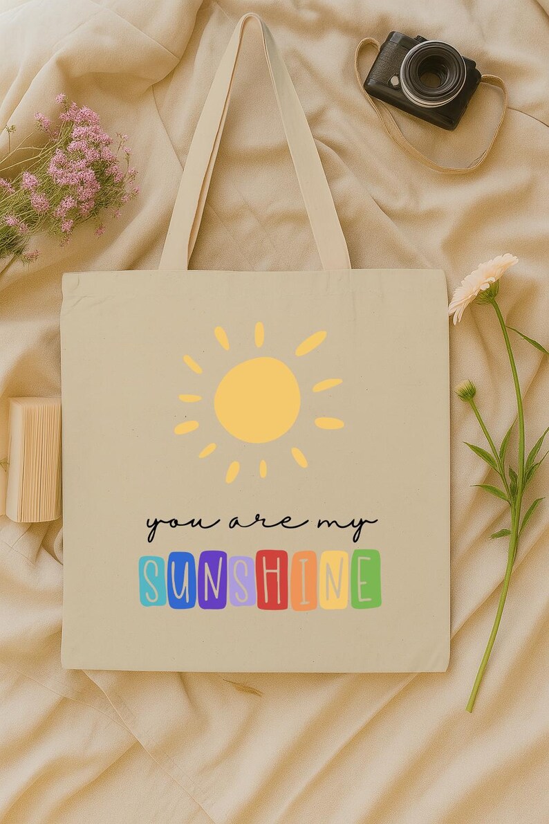 Peut inclure: Sac fourre-tout beige avec un soleil jaune et le texte "you are my sunshine" en lettres color&eacute;es. Le sac a de longues anses et est fait d'un mat&eacute;riau semblable &agrave; de la toile. Un appareil photo noir et des fleurs sont en arri&egrave;re-plan.