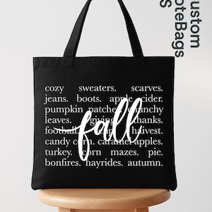 May include: Black tote bag with white text listing fall-themed words like "cozy," "sweaters," and "pumpkin patches." The word "fall" is written in a cursive font. The bag has black handles and is sitting on a wooden stool.