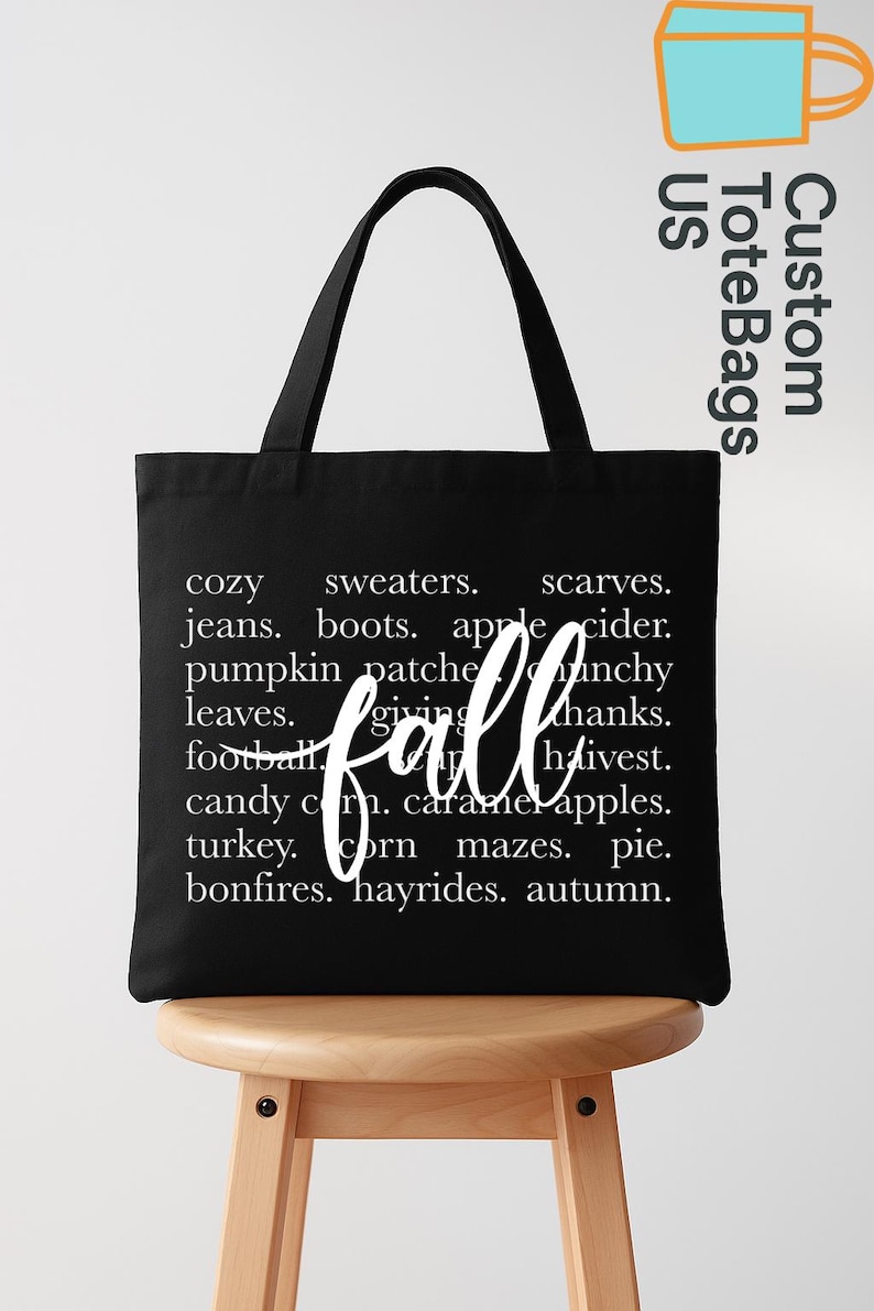 May include: Black tote bag with white text listing fall-themed words like "cozy," "sweaters," and "pumpkin patches." The word "fall" is written in a cursive font. The bag has black handles and is sitting on a wooden stool.