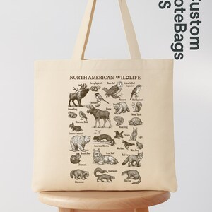 May include: A cream-coloured tote bag displaying a detailed illustration of North American wildlife, including animals such as bears, owls, and foxes. The bag features the text "NORTH AMERICAN WILDLIFE" and "Custom ToteBags US".