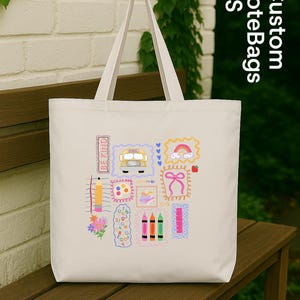 May include: Cream-coloured tote bag featuring colourful school-themed illustrations. The design includes a yellow school bus, a rainbow, crayons, and the words "Be Kind". The bag has two handles and is on a wooden bench.