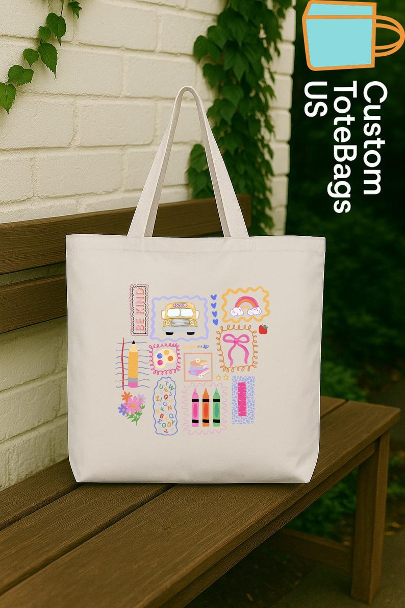 May include: Cream-coloured tote bag featuring colourful school-themed illustrations. The design includes a yellow school bus, a rainbow, crayons, and the words "Be Kind". The bag has two handles and is on a wooden bench.