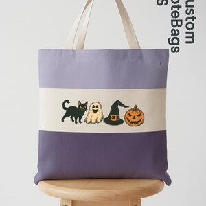 May include: A purple and cream tote bag with a Halloween-themed design. The bag features a black cat, ghost, witch hat, and jack-o'-lantern. The text "Custom Tote Bags US" is printed on the side. The bag has cream-colored handles.