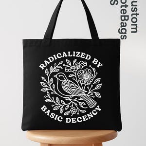May include: Black tote bag with a white graphic of a bird surrounded by flowers and leaves. Text on the bag reads "RADICALIZED BY BASIC DECENCY". The bag has two black handles and is sitting on a wooden stool.