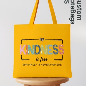 May include: A bright yellow tote bag with the word "KINDNESS" in colorful letters. The phrase "is free" and "SPRINKLE IT EVERYWHERE" are also printed on the bag. The bag has long yellow handles and is sitting on a wooden stool.