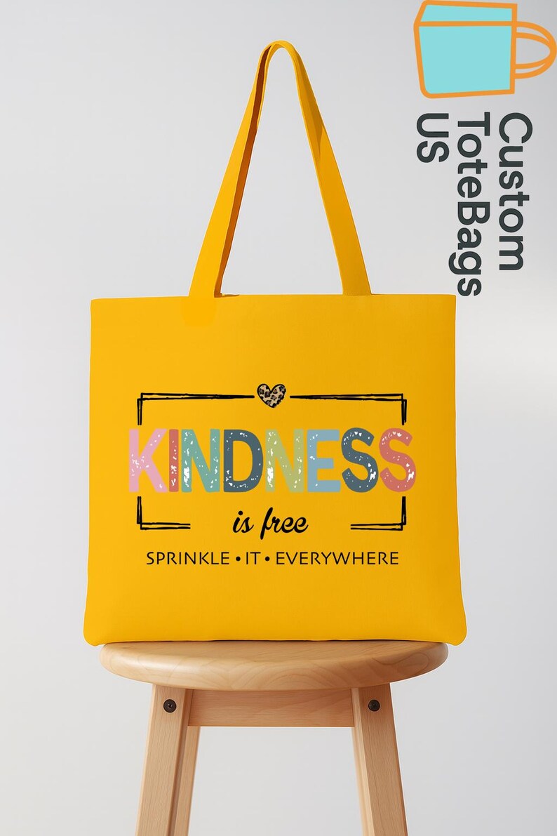 May include: A bright yellow tote bag with the word "KINDNESS" in colorful letters. The phrase "is free" and "SPRINKLE IT EVERYWHERE" are also printed on the bag. The bag has long yellow handles and is sitting on a wooden stool.