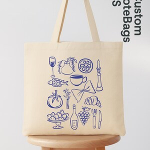 May include: Cream-colored tote bag with a blue line drawing of various food items, including a wine glass, fish, and grapes. The bag has long handles and the text "Custom Tote Bags US" in blue.