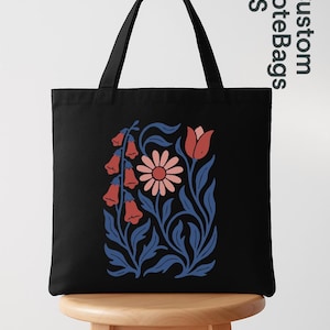 Peut inclure: Sac fourre-tout noir avec un motif floral de fleurs en clochettes rouges, une marguerite rose et des feuilles bleues. Le sac a des anses noires et est pos&eacute; sur un tabouret en bois. Le texte "Custom ToteBags US" est imprim&eacute; sur le c&ocirc;t&eacute;.