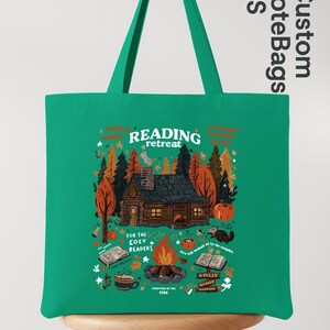 May include: A green tote bag featuring a cabin scene with a campfire and the words "Reading Retreat." The design includes pumpkins, trees, and books, with the text "For the Cozy Readers."