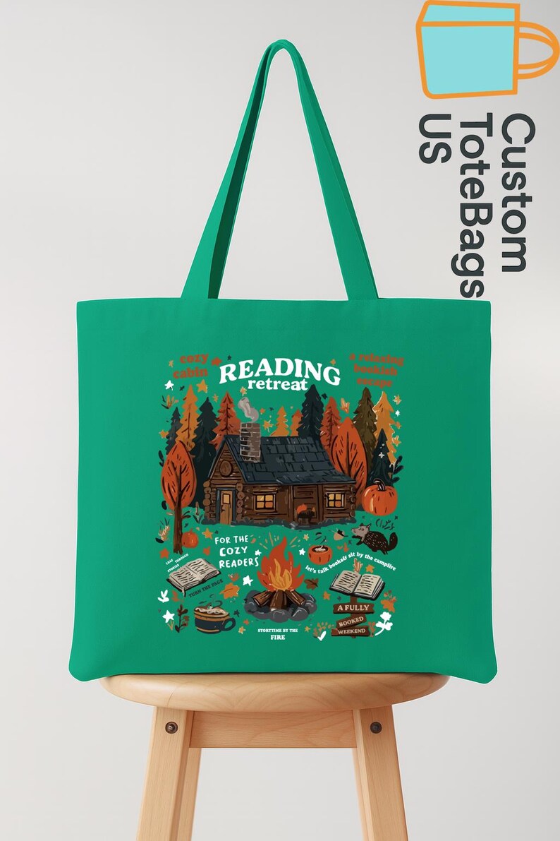May include: A green tote bag featuring a cabin scene with a campfire and the words "Reading Retreat." The design includes pumpkins, trees, and books, with the text "For the Cozy Readers."