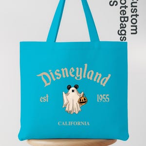 May include: A turquoise tote bag with the Disneyland logo, a ghost character holding a pumpkin, and the text "est 1955 CALIFORNIA". The bag has matching turquoise handles and is displayed on a wooden stool.