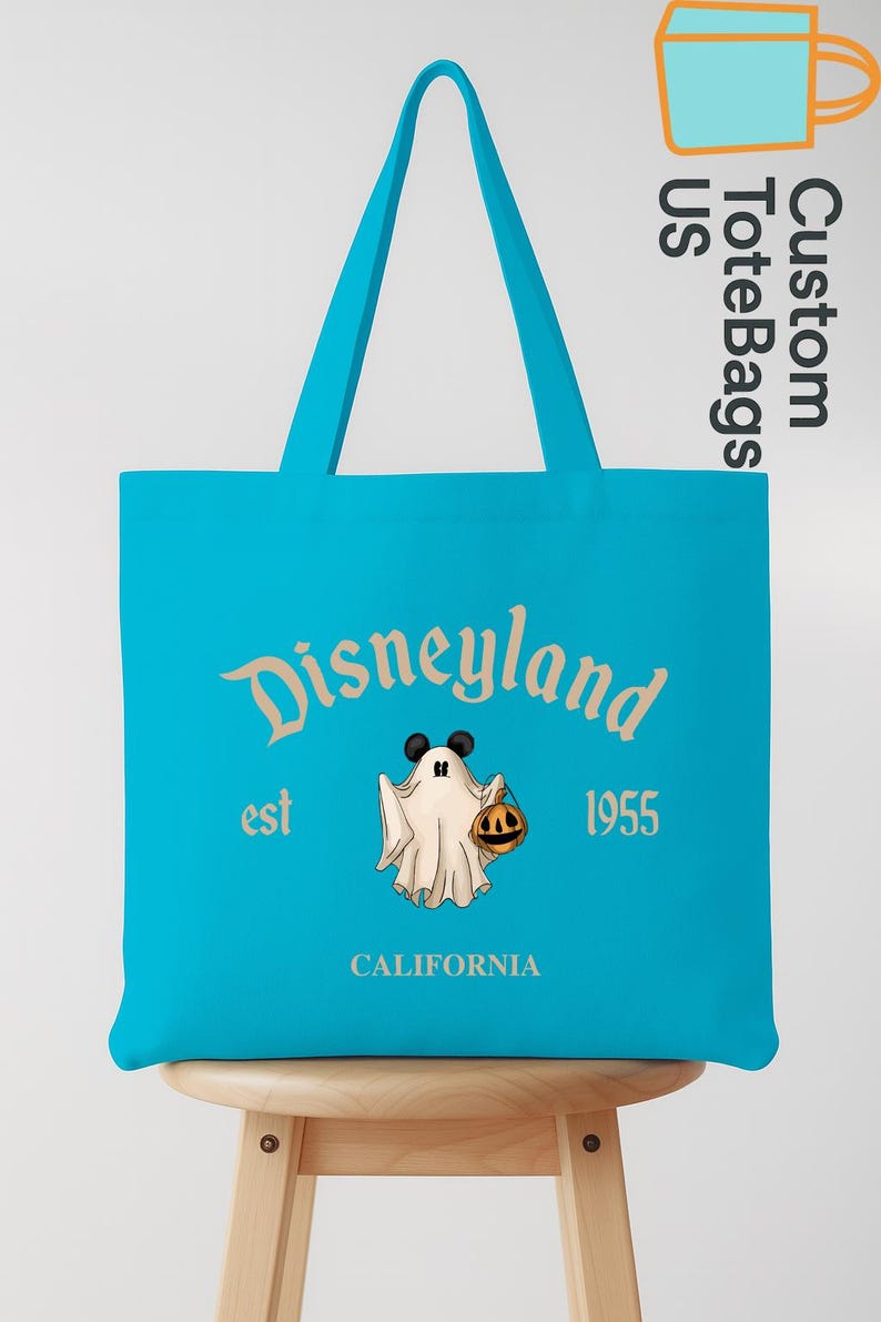 May include: A turquoise tote bag with the Disneyland logo, a ghost character holding a pumpkin, and the text "est 1955 CALIFORNIA". The bag has matching turquoise handles and is displayed on a wooden stool.