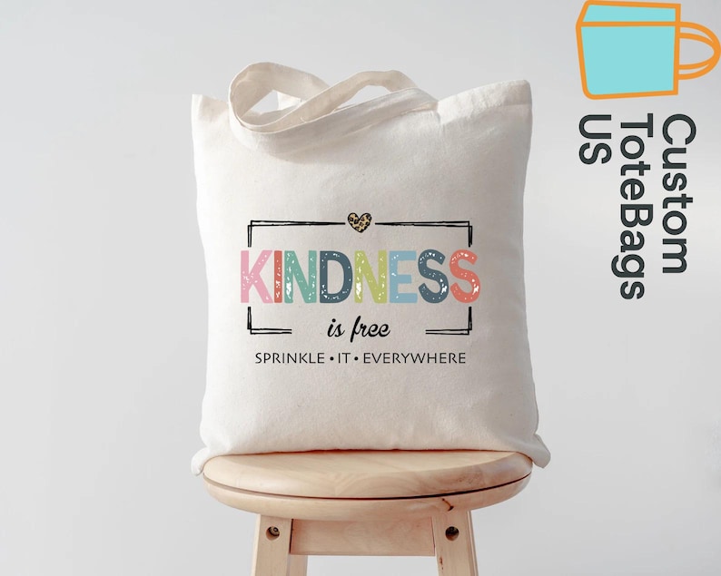 May include: A white canvas tote bag with the words "KINDNESS is free" in colorful letters. The bag has a black rectangular border and a leopard print heart. The phrase "SPRINKLE IT EVERYWHERE" is below the main text. The bag has cream-colored handles.