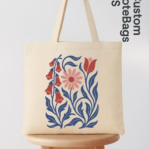Peut inclure: Sac fourre-tout cr&egrave;me avec un motif floral de fleurs en clochettes rouges, une marguerite rose et des feuilles bleues. Le sac a de longues anses et le texte "Custom Tote Bags US".