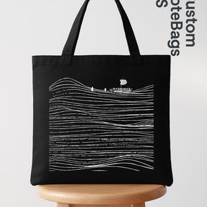 Puede incluir: Bolso tote negro con un dibujo lineal blanco de un barco navegando sobre l&iacute;neas onduladas. El bolso tiene dos asas negras y una forma rectangular. Las palabras "Custom Tote Bags US" est&aacute;n impresas en la esquina superior derecha.