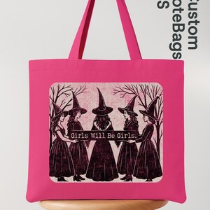 May include: A vibrant pink tote bag with a graphic of five witches in black dresses and hats, standing before bare trees. The text "Girls Will Be Girls" is below the image. The bag has long handles and is on a wooden stool.