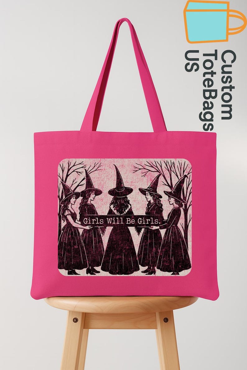 May include: A vibrant pink tote bag with a graphic of five witches in black dresses and hats, standing before bare trees. The text "Girls Will Be Girls" is below the image. The bag has long handles and is on a wooden stool.