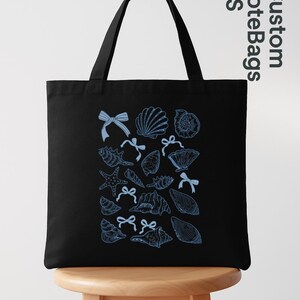 K&ouml;nnte beinhalten: Schwarze Tragetasche mit blauem Muschel- und Schleifenmuster. Die Tasche hat zwei schwarze Griffe und wird auf einem Holzschemel gezeigt. Der Text "Custom ToteBags US" ist oben rechts sichtbar.