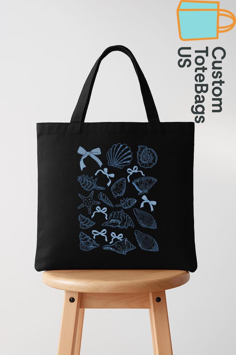 K&ouml;nnte beinhalten: Schwarze Tragetasche mit blauem Muschel- und Schleifenmuster. Die Tasche hat zwei schwarze Griffe und wird auf einem Holzschemel gezeigt. Der Text "Custom ToteBags US" ist oben rechts sichtbar.