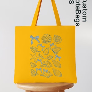 K&ouml;nnte beinhalten: Gelbe Tragetasche mit blauem Muschel- und Schleifenmuster. Die Tasche hat lange Henkel und steht auf einem Holzschemel. Der Text "Custom Tote Bags US" ist auf der Seite aufgedruckt.