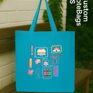 May include: A turquoise tote bag with school-themed illustrations, including a school bus, rainbow, pencil, and crayons. The text "BE KIND" is printed vertically. The bag has matching turquoise handles.