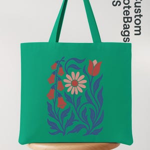 Peut inclure: Un sac fourre-tout vert avec un motif floral comprenant des fleurs rouges en forme de cloche, une marguerite rose et des feuilles bleues. Le sac a des poign&eacute;es vertes et le texte "Custom Tote Bags US".