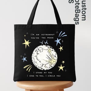 Peut inclure: Sac fourre-tout noir avec un design fantaisiste représentant une lune, des étoiles et le texte "I'm an astronaut, you're the moon." Le sac présente une illustration simple mais charmante, idéale pour transporter des livres ou des courses.