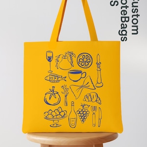 May include: Yellow tote bag with blue line art of food items like a wine glass, fish, tomato, and pasta. The bag has long handles and the text "Custom Tote Bags US" in the upper right corner.