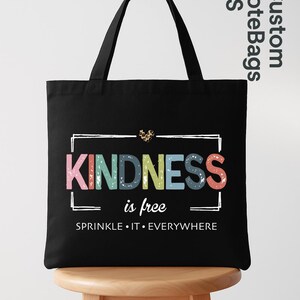 May include: Black tote bag with the words "KINDNESS is free" in colorful letters. The bag has a white rectangular border around the text and a leopard print heart above the word "KINDNESS". The phrase "SPRINKLE IT EVERYWHERE" is below the main text.