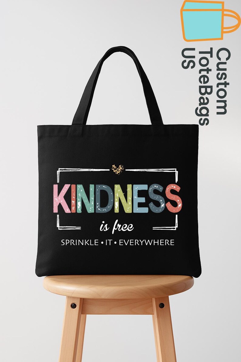May include: Black tote bag with the words "KINDNESS is free" in colorful letters. The bag has a white rectangular border around the text and a leopard print heart above the word "KINDNESS". The phrase "SPRINKLE IT EVERYWHERE" is below the main text.