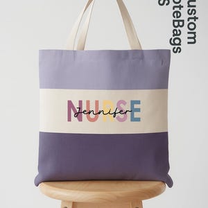 May include: A tote bag with a lavender and purple color block design. The bag features the word "NURSE" in colorful letters, with the name "Jennifer" below. The bag has off-white handles and is displayed on a wooden stool.