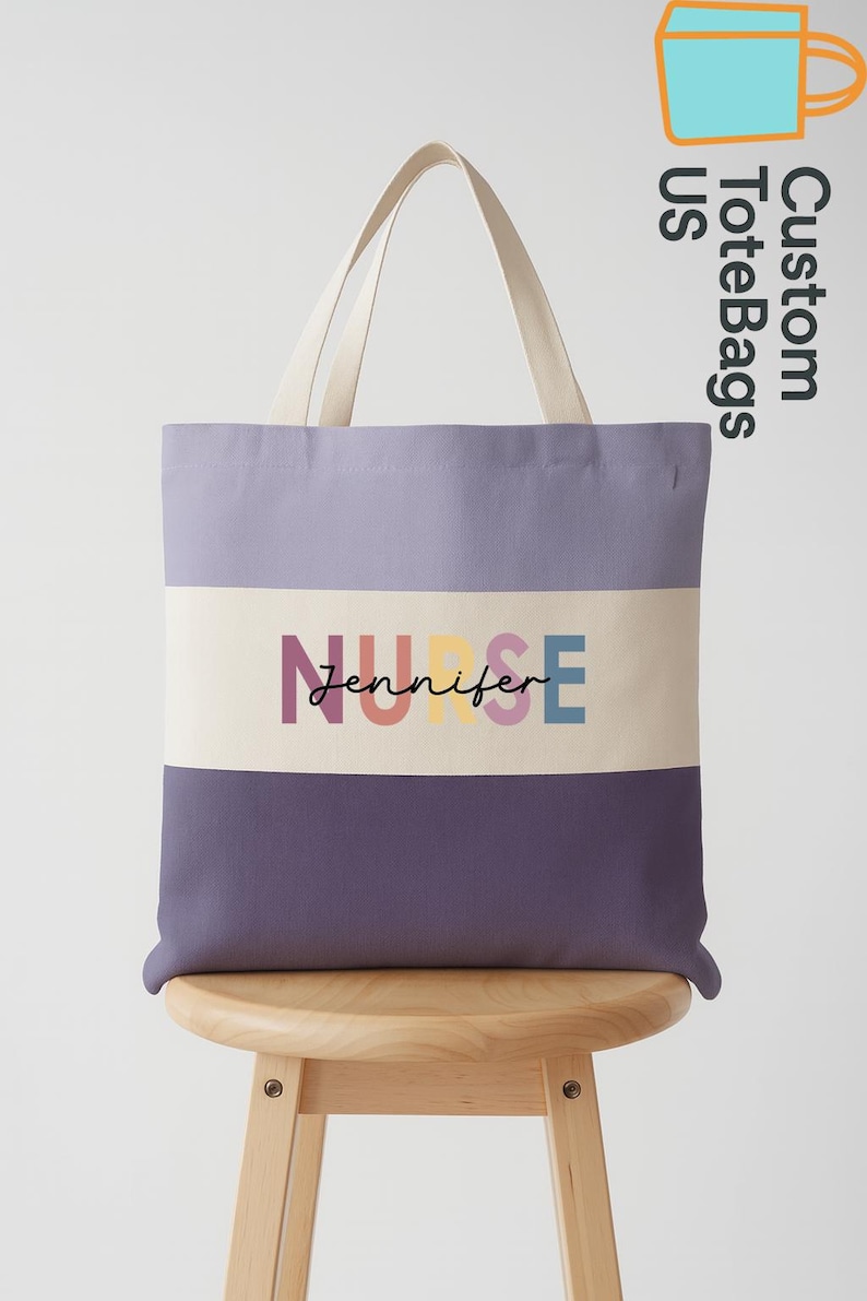 May include: A tote bag with a lavender and purple color block design. The bag features the word "NURSE" in colorful letters, with the name "Jennifer" below. The bag has off-white handles and is displayed on a wooden stool.