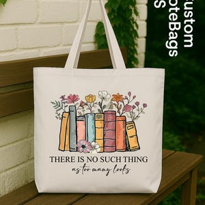 May include: A white tote bag featuring a colorful illustration of books with flowers on top. The text "THERE IS NO SUCH THING as too many books" is printed below the books. The bag has a simple design with two handles.