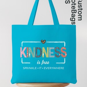 May include: A turquoise tote bag with the words "KINDNESS is free" in colorful letters. The bag has a leopard print heart above the text and the phrase "SPRINKLE IT EVERYWHERE" below. The bag is sitting on a wooden stool.