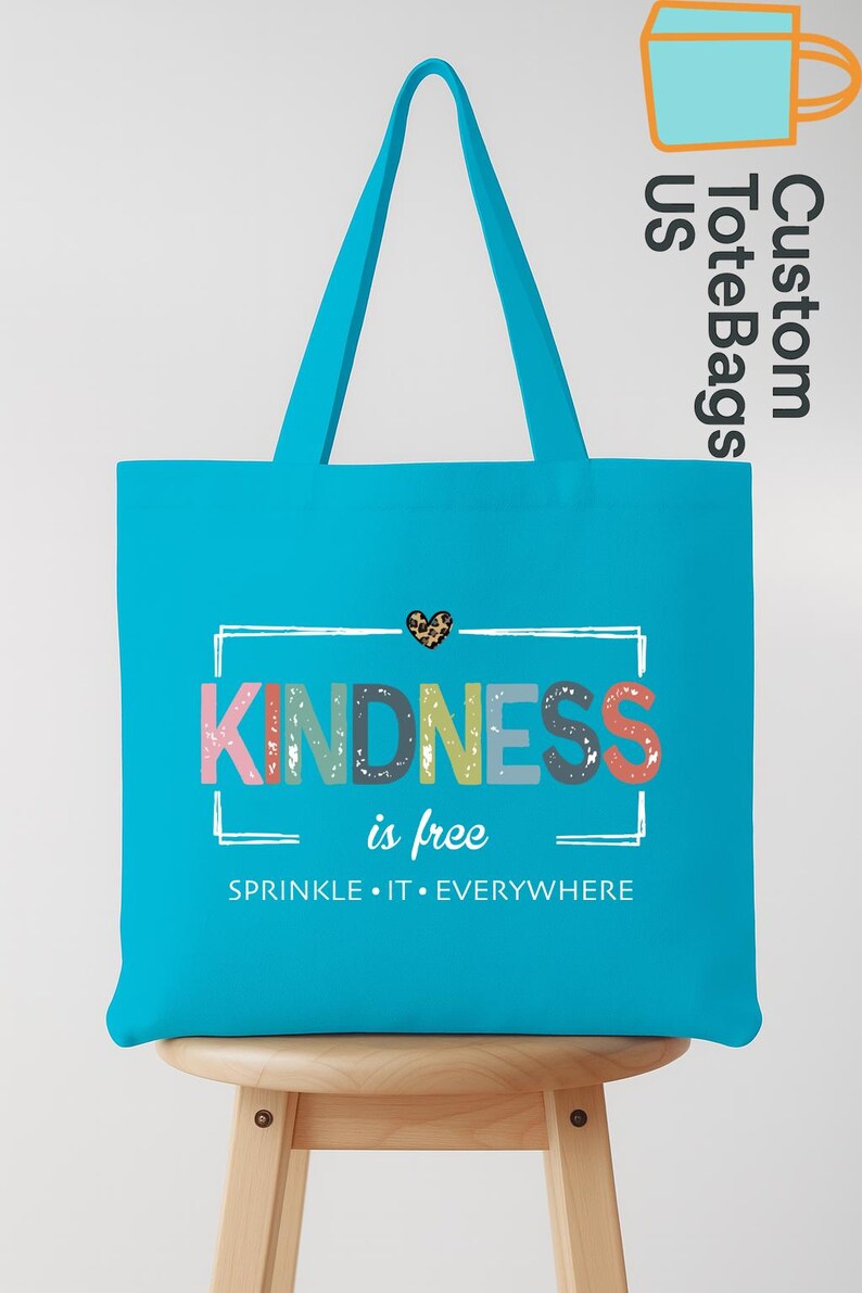 May include: A turquoise tote bag with the words "KINDNESS is free" in colorful letters. The bag has a leopard print heart above the text and the phrase "SPRINKLE IT EVERYWHERE" below. The bag is sitting on a wooden stool.