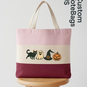 May include: A striped tote bag with a pink, beige, and burgundy color scheme. The bag features a Halloween-themed graphic of a black cat, ghost, witch hat, and jack-o'-lantern. The text "Custom Tote Bags US" is printed on the side.